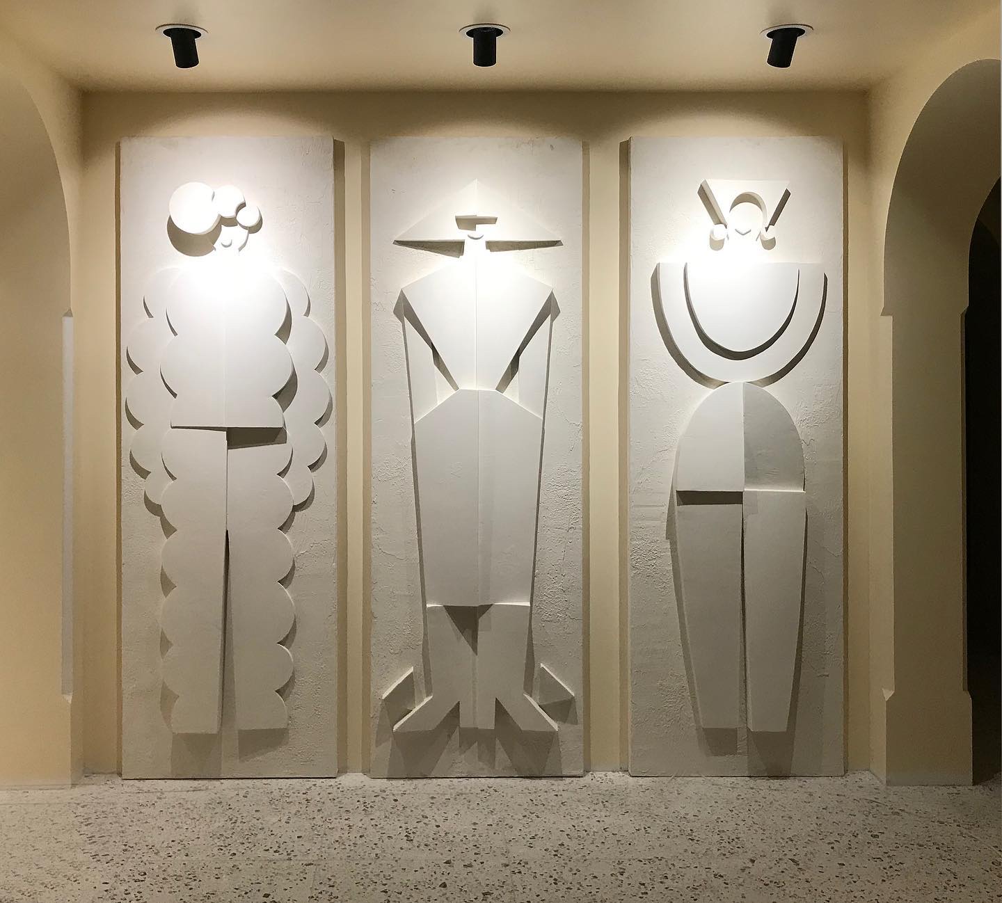 Three tall white relief panels of stylised figures, lit from above.